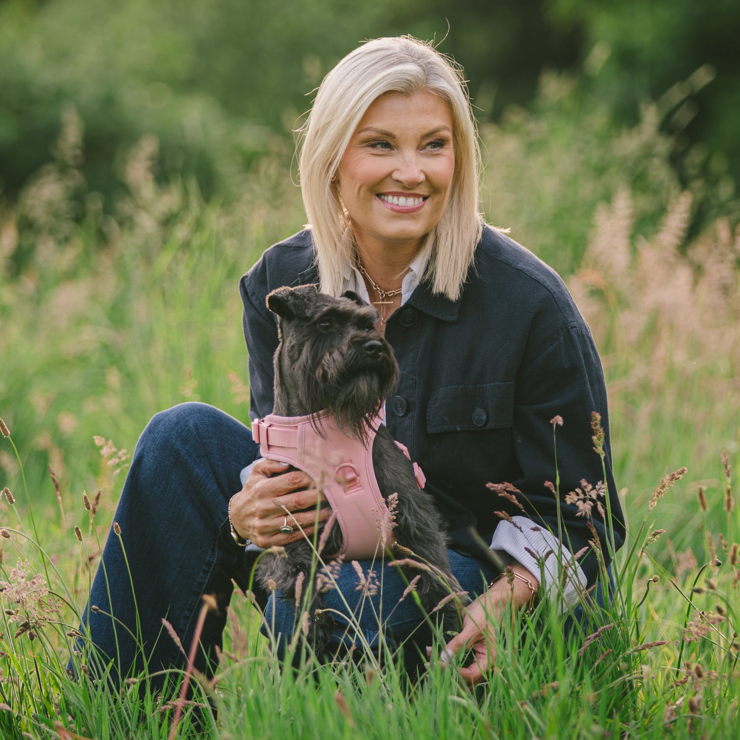 Darcy and Ted founder, Gillian, in a grassy field holding a small black dog, Darcy wearing a pink harness.