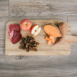 Wooden cutting board with raw beef, apples, carrots, and a sweet potato on a wooden floor.