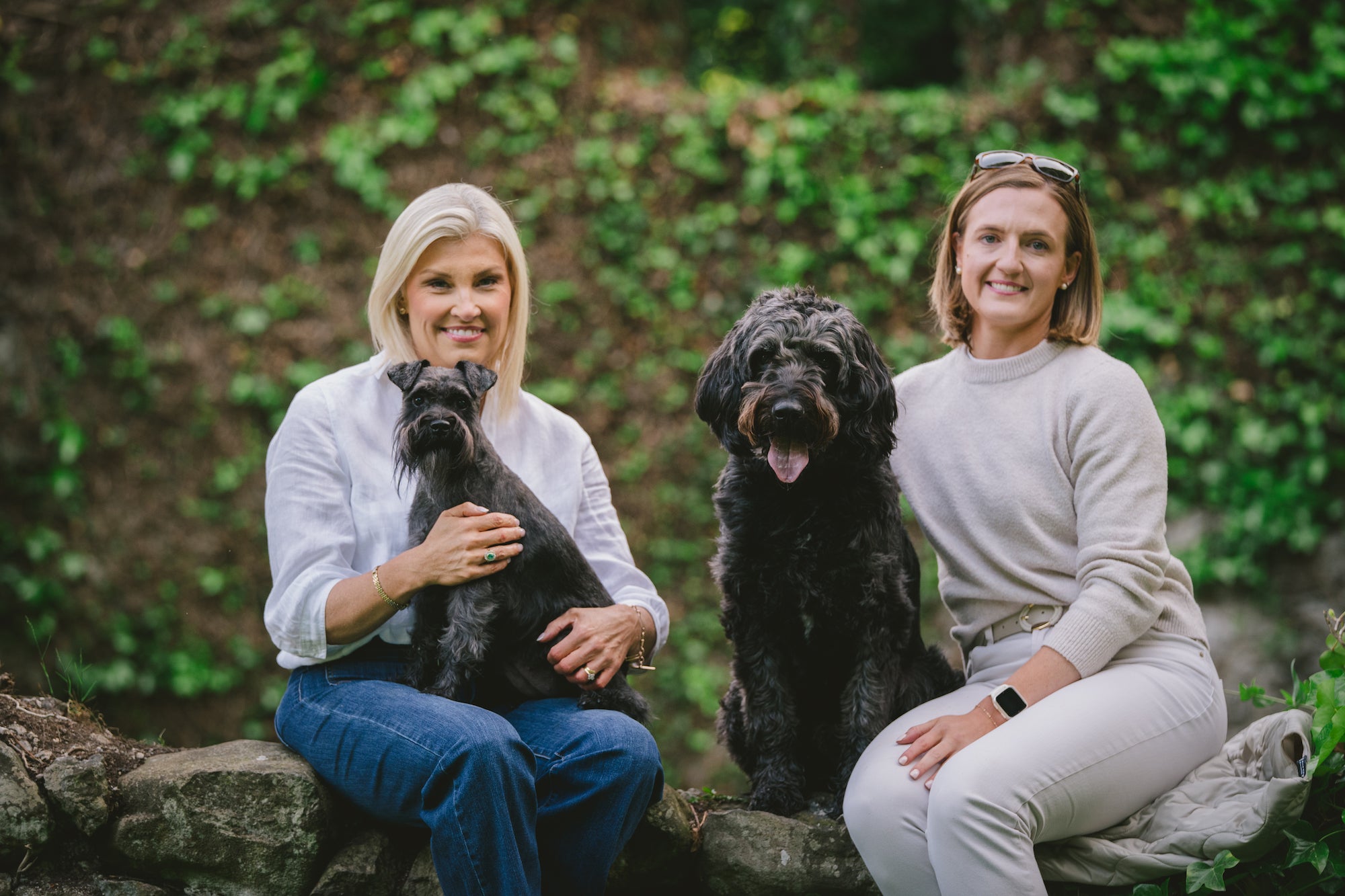 Darcy and Ted's founders, Joanne and Gillian, sitting on a stone wall with their two dogs, Darcy and Ted, surrounded by greenery.