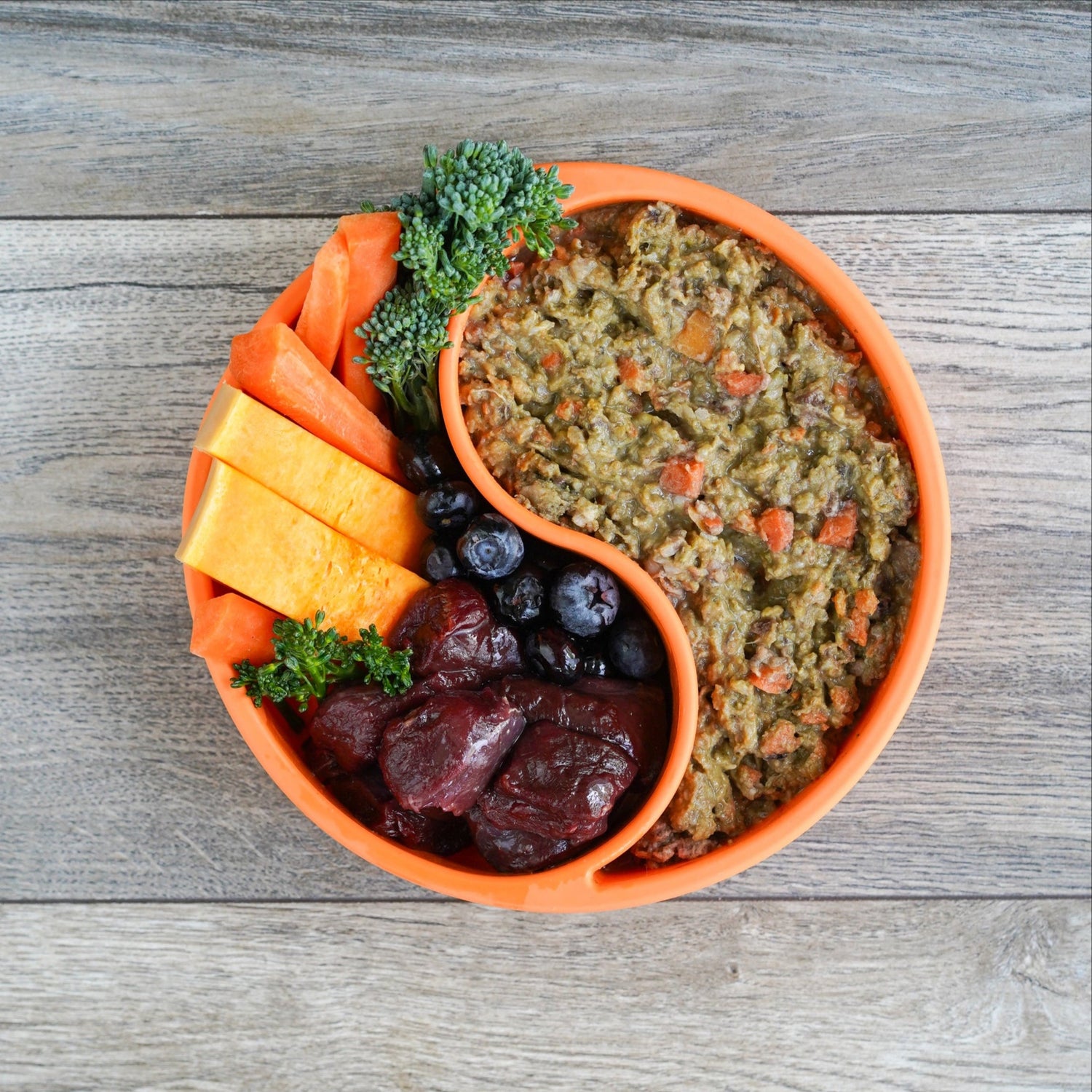 Dish with duck delight raw dog food, including raw duck, and vegetables, on a wooden surface.