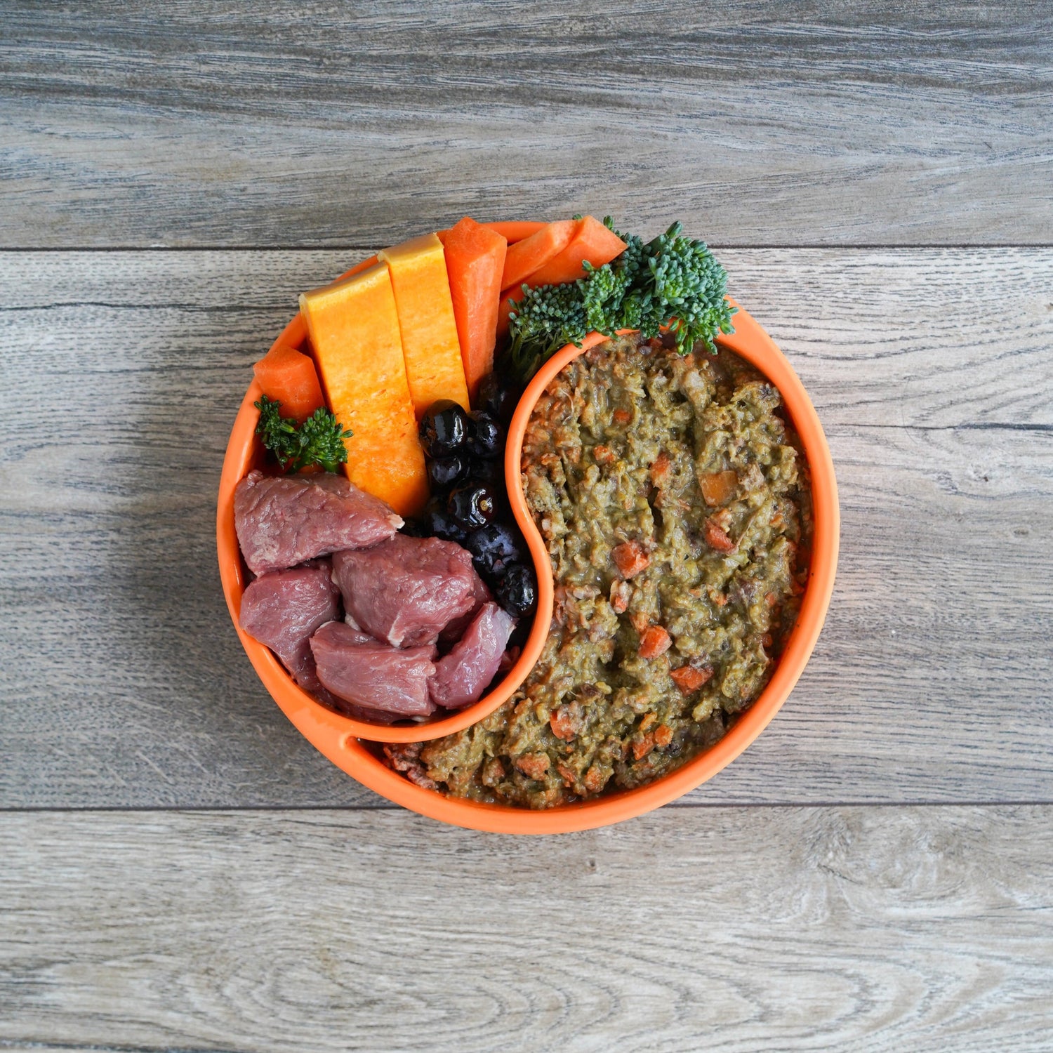 Pet food bowl with loaded lamb, raw meat, carrots, and broccoli on a wooden surface